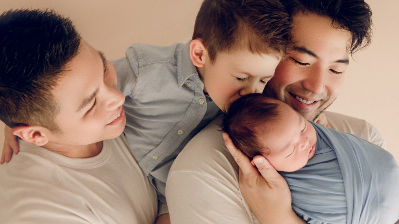 A baby swaddled in a lightweight muslum swaddle cloth being held by his two daddies. 